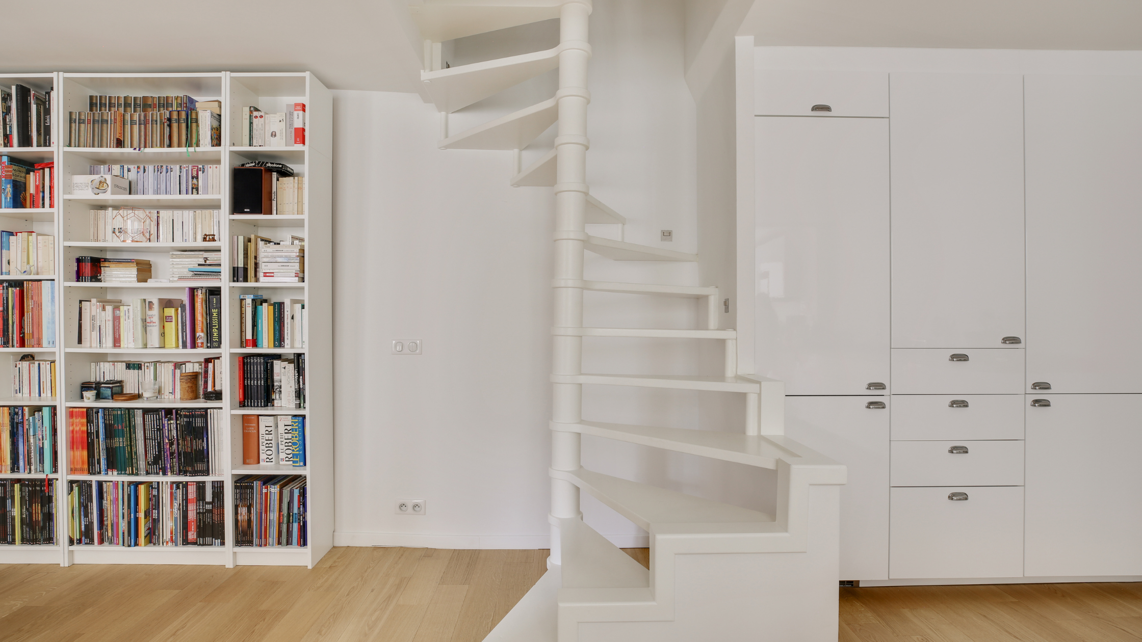 image d'un intéireur blanc ou l'on voit au centre un escalier en colimaçon blanc design sans garde corps, à gauche on aperçoit une bibliothèque ikea et à droite des placards de cuisine ikea.c'est le travail d'un architecte d'intérieur à distance